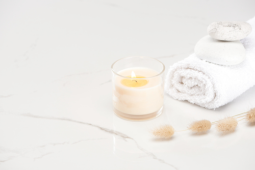 fluffy bunny tail grass near burning white candle in glass and rolled towel with stones on marble white surface