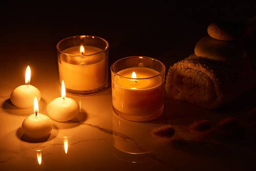 burning candles in glass glowing in dark near rolled towel on marble surface