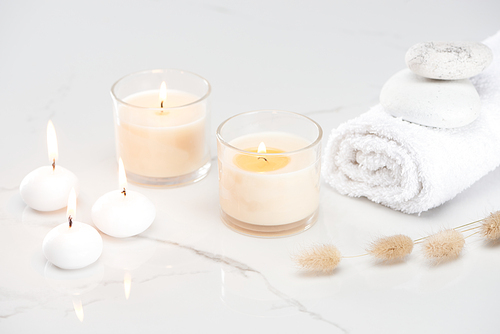fluffy bunny tail grass near burning white candles in glass and rolled towel with stones on marble white surface