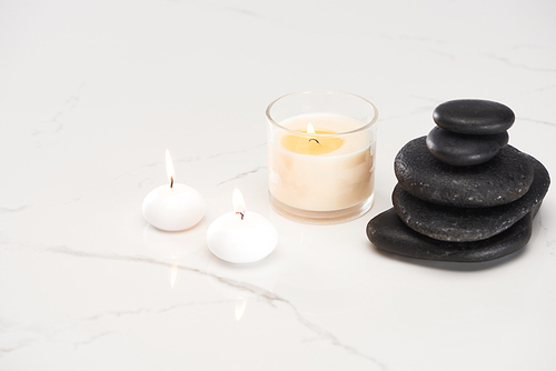 burning white candles in glass near spa stones and rolled towel on marble white surface