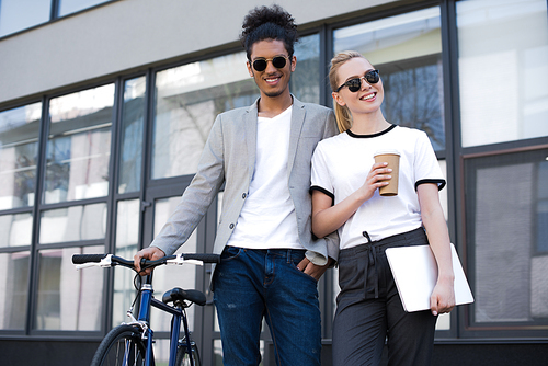 cheerful young multiethnic couple standing with laptop, coffee to go and bicycle on street