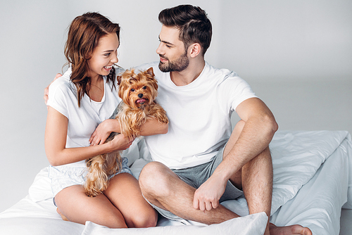 smiling couple in love with yorkshire terrier resting on bed together isolated on grey