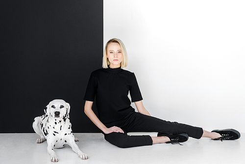 attractive stylish blonde woman in black clothes sitting near black and white wall with dalmatian dog