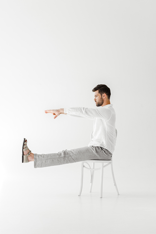 profile of male model in linen clothes sitting on chair with outstretched legs and hands isolated on grey background