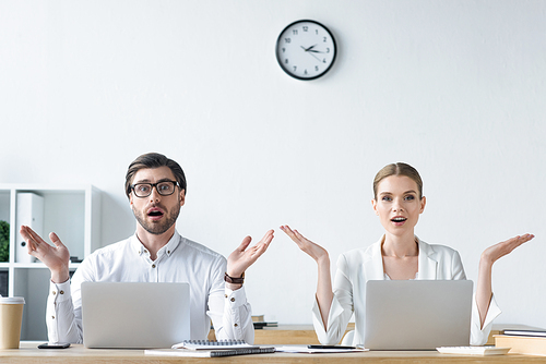 surprised young managers raising hands while working with laptops together at office