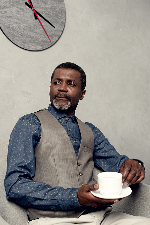 serious african american man in waistcoat sitting in armchair with cup of coffee