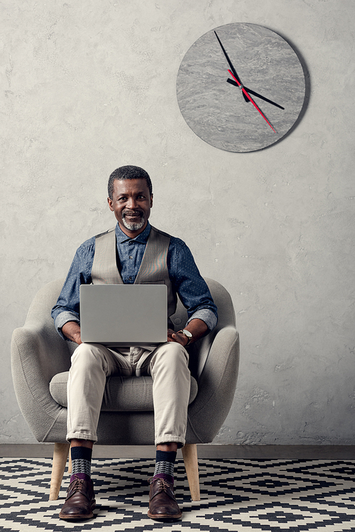 smiling african american businessman using laptop in armchair in office with clock on wall