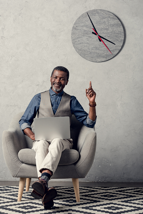stylish african american businessman pointing up and using laptop in office with clock on wall