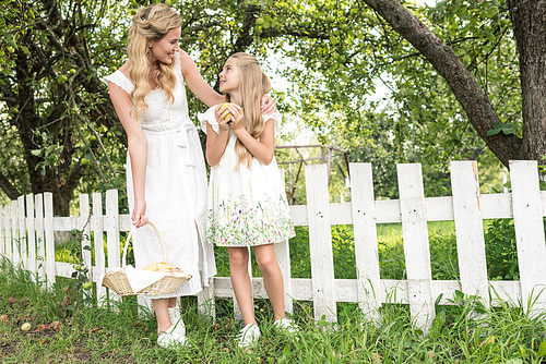 blonde mother and daughter with fruits in wicker basket posing near white fence in garden