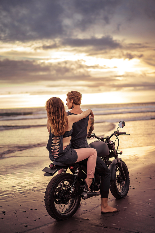 back view of boyfriend and girlfriend sitting on motorbike on seashore during beautiful sunrise