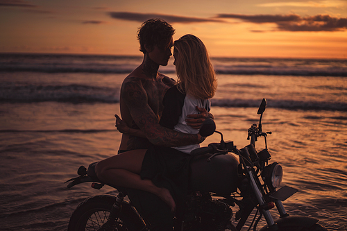 passionate boyfriend and girlfriend hugging on motorbike at beach during sunset