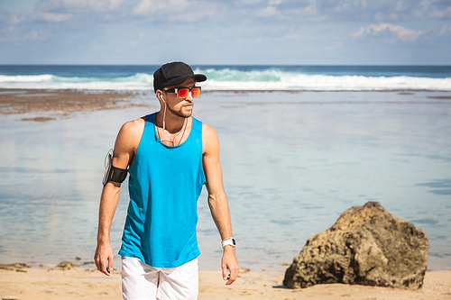athletic man in sunglasses with armband walking on beach near sea, Bali, Indonesia