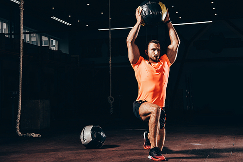 handsome athletic bodybuilder performing lunge with medicine ball overhead in dark gym