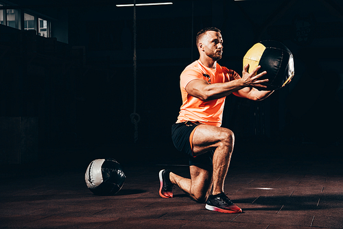 handsome athletic sportsman performing lunge with medicine ball in dark gym