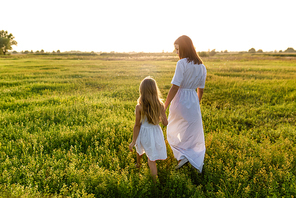 mother and daughter holding hands and walking by green meadow with sunset sky on background