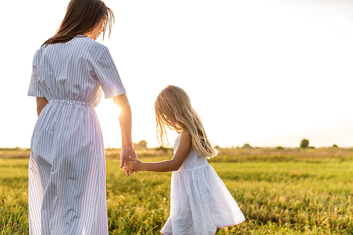 rear view of mother and daughter in white dresses holding hands in green meadow on sunset