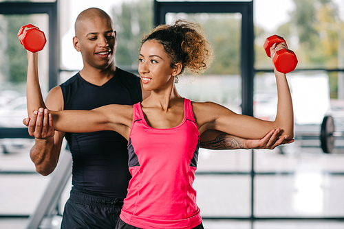 smiling african american male trainer helping sportswoman to exercising with dumbbells at gym