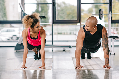 smiling african american couple of athletes looking at each other and doing plank on fitness mats at gym