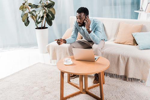 handsome african american freelancer sitting on couch and talking on smartphone