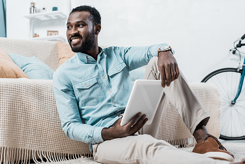 african american man holding digital tablet in hands and looking away in living room