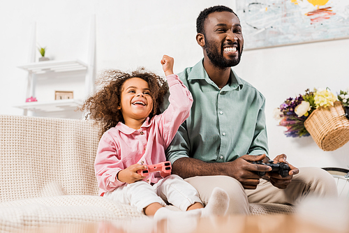 african american dad laughing with daughter and playing video game in living room