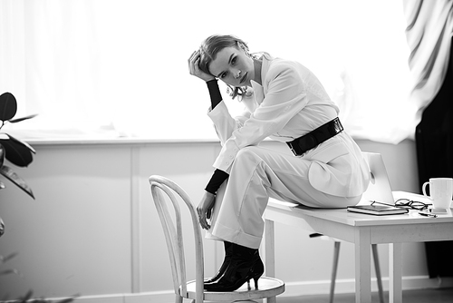 black and white photo of stylish businesswoman in formal wear sitting on desk and  at workplace