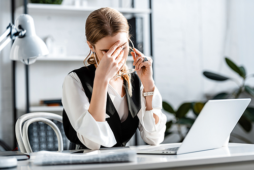 selective focus of businesswoman in formal wear sitting at computer desk, touching forehead and suffering from headache at workplace