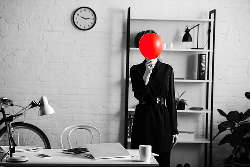 black and white of businesswoman in black clothes standing near table and shelves and hiding face behind red balloon