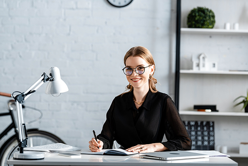 beautiful businesswoman in black clothes and glasses smiling and writing in note