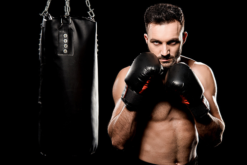 athlete in boxing gloves standing in boxing pose near punching bag isolated on black