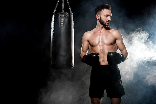 pensive sportsman in boxing gloves standing near punching bag on black with smoke