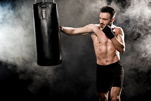 shortless athletic boxer exercising with punching bag on black with smoke