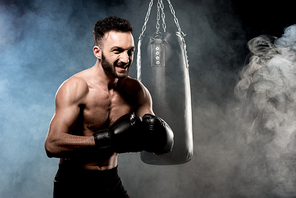 angry boxer standing in boxing pose near punching bag on black with smoke