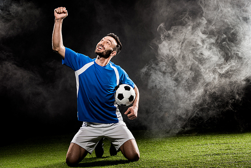 cheerful football player celebrating victory and holding ball on black with smoke