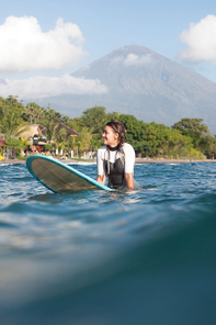 smiling woman sitting on surf board in ocean, coastline on background