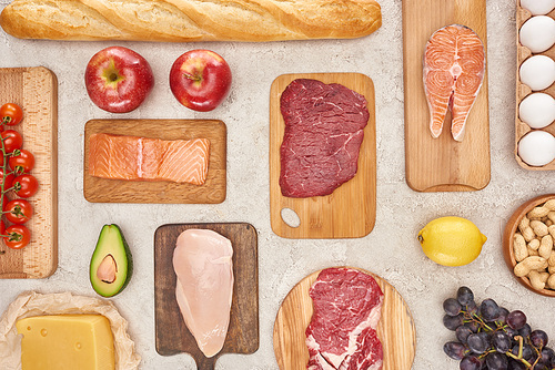 top view of assorted meat, poultry, fish, eggs, fruits, s, cheese and baguette on wooden cutting boards on marble surface