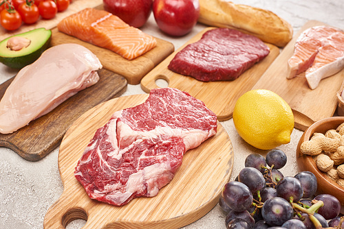 Fresh raw meat, poultry, fish on wooden cutting boards near lemon, grapes, apples, branch of cherry tomatoes, nuts and french baguette on marble surface
