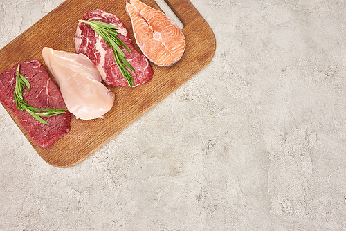 Top view of assorted meat, poultry and fish with rosemary twigs on wooden cutting board on gray marble surface