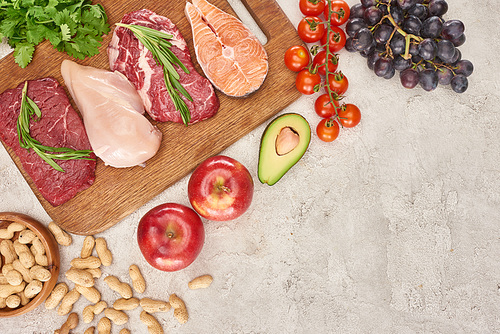 Top view of assorted meat, poultry and fish with greenery on wooden cutting board near apples, grapes, peanuts, cherry tomatoes and half of avocado on gray marble surface