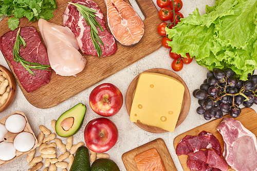 Top view of assorted meat, poultry and fish with greenery and cheese on wooden cutting boards near apples, grapes, peanuts, cherry tomatoes, eggs and avocados on gray marble surface