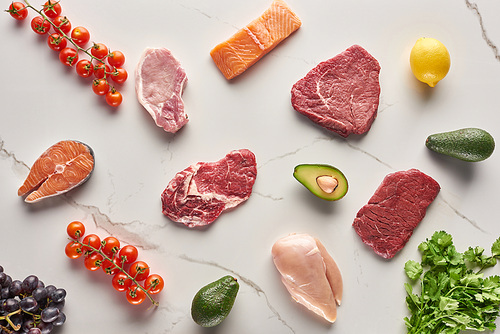 Top view of assorted meat, poultry and fish near parsley, grapes, cherry tomatoes, avocados and lemon on gray marble surface