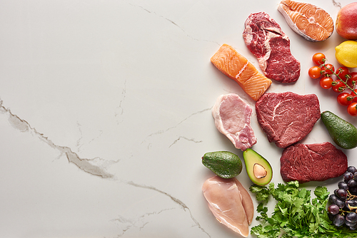 Top view of assorted meat, poultry and fish near parsley, grapes, cherry tomatoes, avocados, apple and lemon on gray marble surface