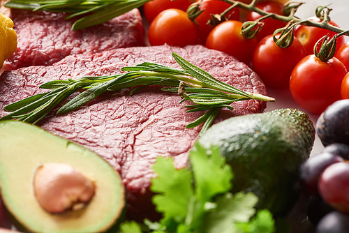 Close up view of raw meat steak with rosemary twig near avocados, branch of cherry tomatoes and parsley