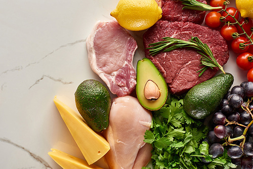 Top view of assorted raw meat and poultry with avocados, lemon, cheese, grapes, branch of tomatoes and greenery twigs