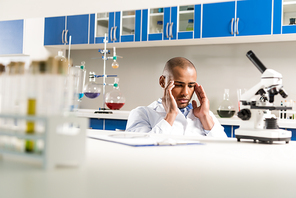 Young pensive technician in lab coat sitting in laboratory and rubbing his temples