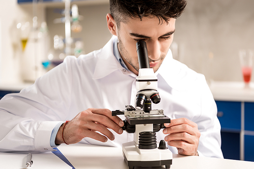 Young lab technician in white coat looking into the microscope in laboratory
