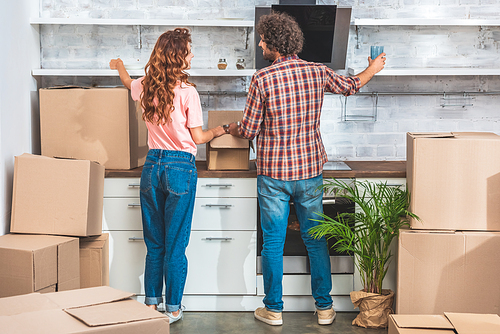 back view of couple unpacking cardboard boxes at new home and putting utensil on shelves