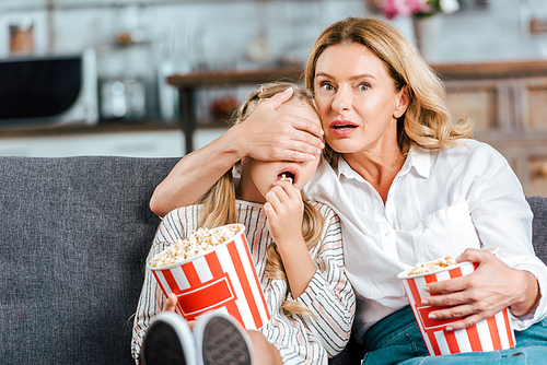 frightened mother and daughter with buckets of popcorn watching scary movie at home