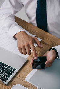 partial view of businessman using smartphone at tabletop with documents and laptop