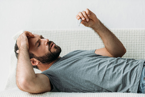 sick bearded man lying on couch and holding thermometer